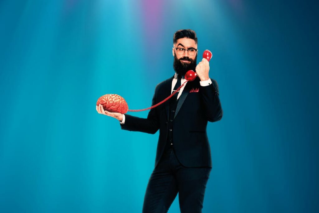 man with dark hair and beard standing on spotlit stage holding a brain in one hand and a telephone in the other