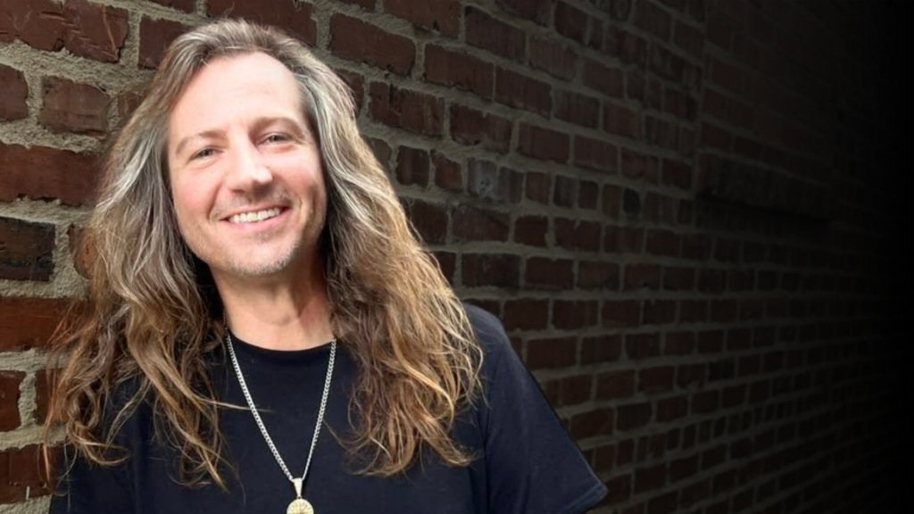 Man with long light brown hair and black t-shirt standing in front of brick wall